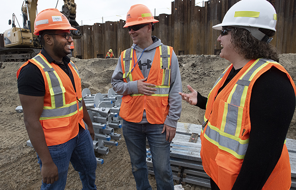 Picture of three people wearing safety vests and hard hats on a work site. Behind them are building materials being used in the project.